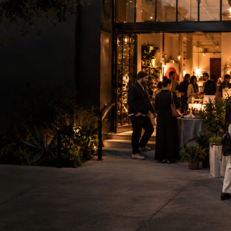 An evening neighborhood retail gathering with local vendor tables leading toward an open storefront.